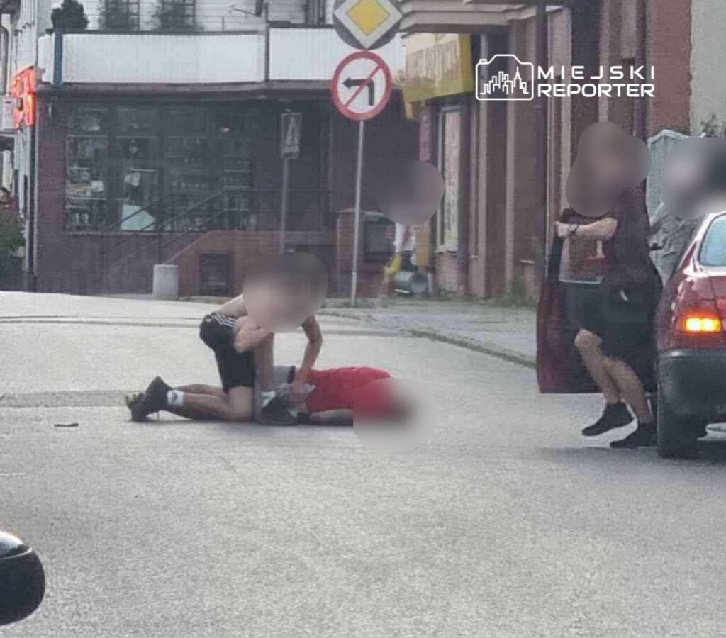 Dwaj mężczyźni na ulicy, jeden leży na ziemi w czerwonym ubraniu, a drugi znajduje się nad nim. W tle widoczny jest sklep i znaki drogowe.