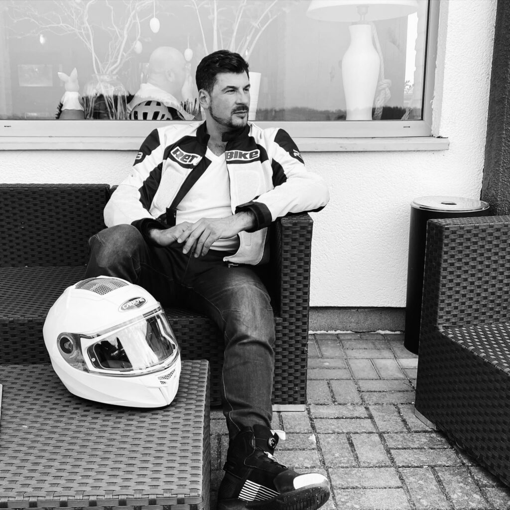 Mężczyzna w kurtce motocyklowej siedzi na rattanowym fotelu obok białego kasku, w tle widać okno z lampą.