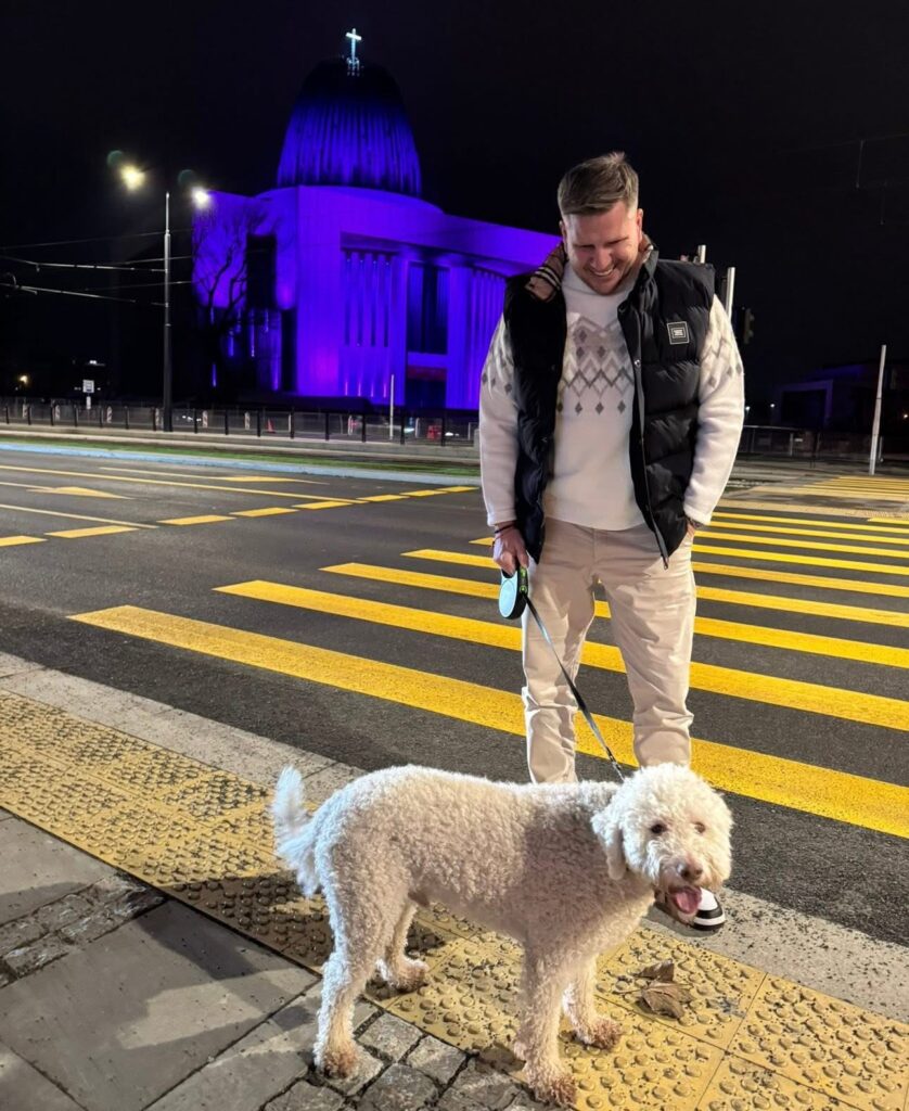 Mężczyzna w czarnej kamizelce trzyma na smyczy białego psa, stojąc na przejściu dla pieszych w nocy. W tle oświetlony budynek.