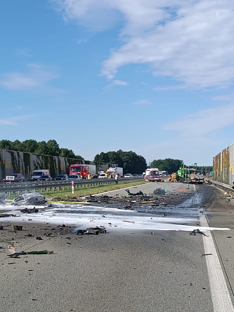Zniszczona nawierzchnia drogi z fragmentami wraku pojazdu i gaśnicą, w tle pojazdy ratunkowe i ruch samochodowy.
