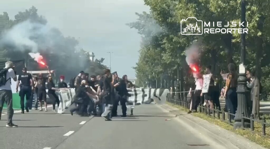 Grupa demonstrantów z transparentem blokuje ruch na ulicy, podczas gdy niektórzy z nich trzymają racę i krzyczą.