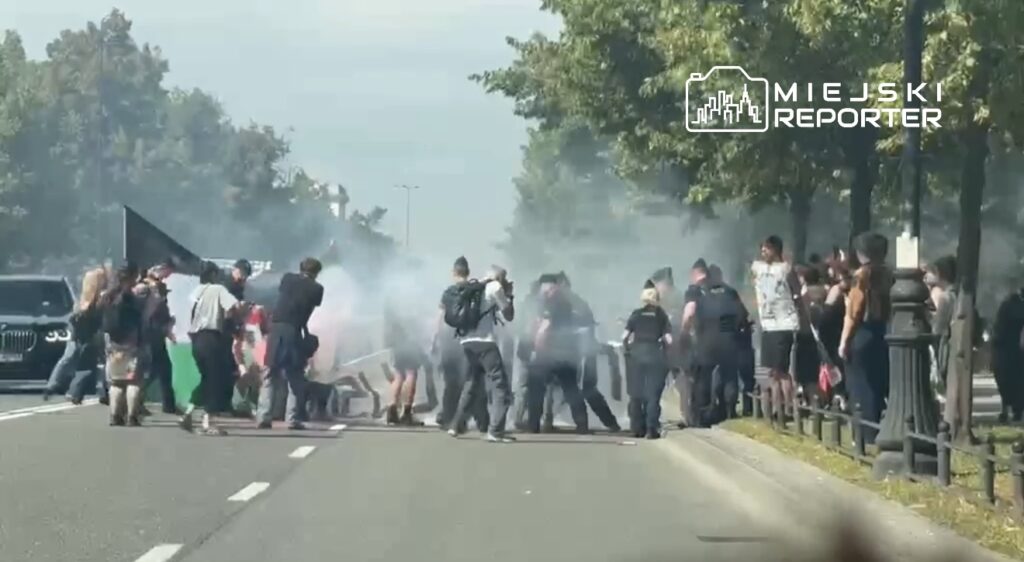 Grupa demonstrantów blokuje drogę, unosząc flagi i wytwarzając dym, podczas gdy policja interweniuje w tle.