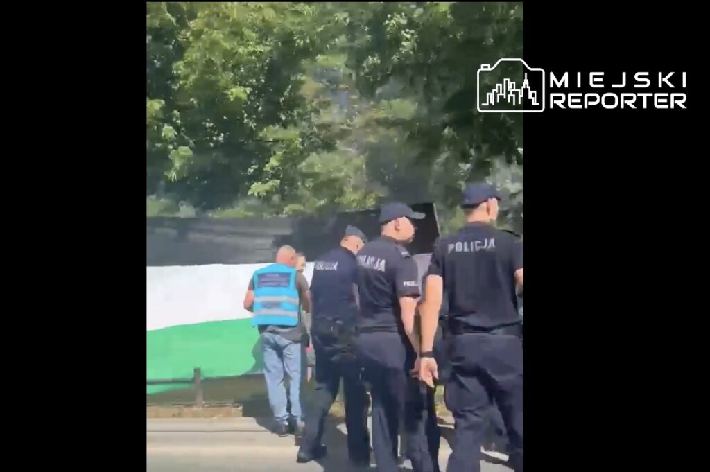 Grupa policjantów w mundurach idzie wzdłuż ogrodzenia, a jeden z mężczyzn w kamizelce rozmawia z nimi.