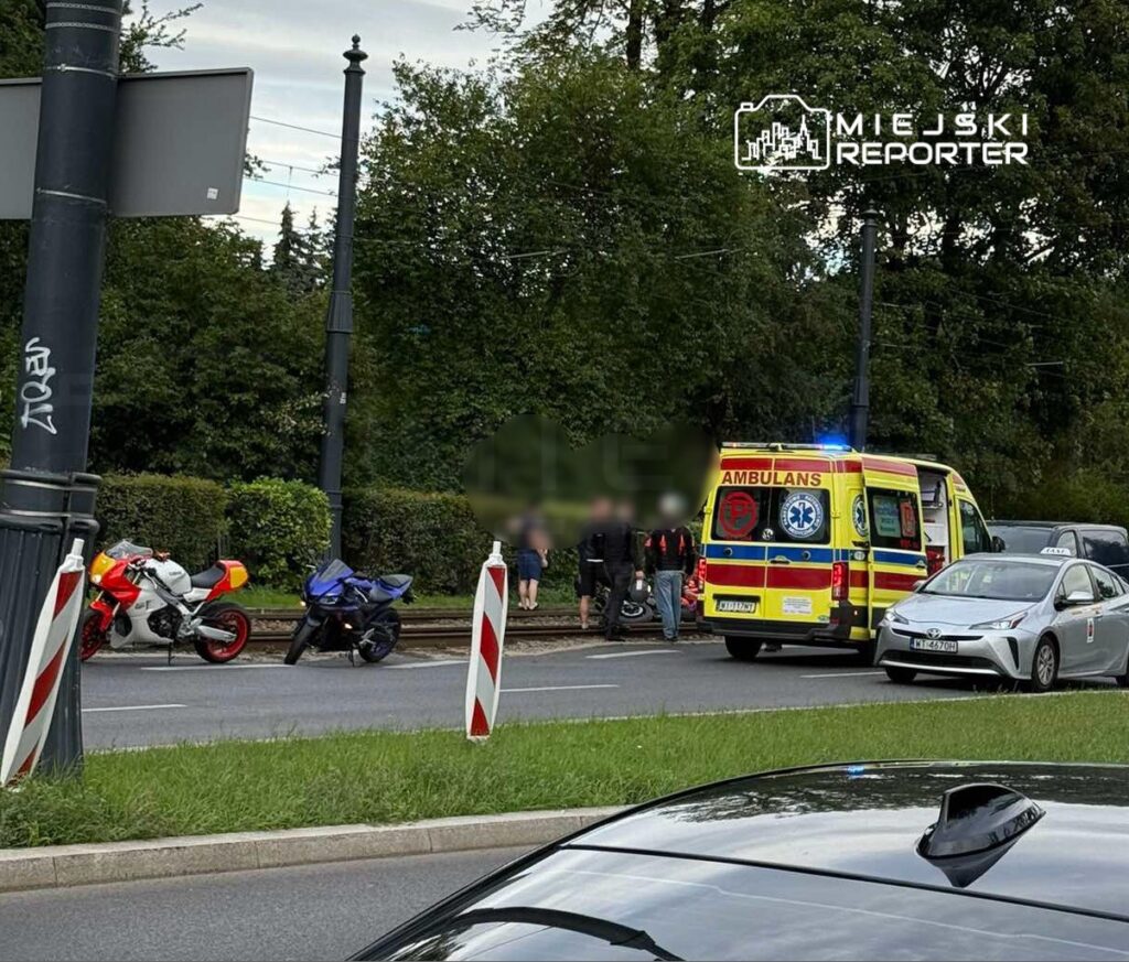 Na ulicy zatrzymane są dwa motocykle, a obok nich stoi karetka pogotowia. Grupa osób obserwuje sytuację.