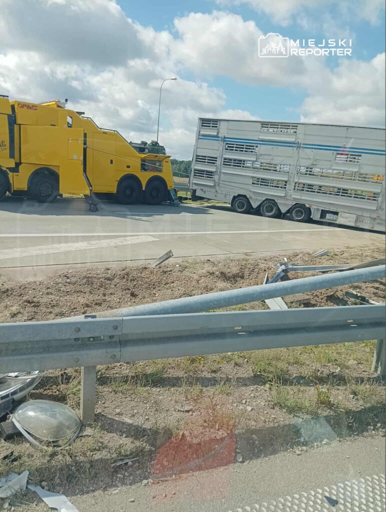 Żółty ciągnik siodłowy holuje przewróconą naczepę na poboczu drogi, obok uszkodzone barierki ochronne.