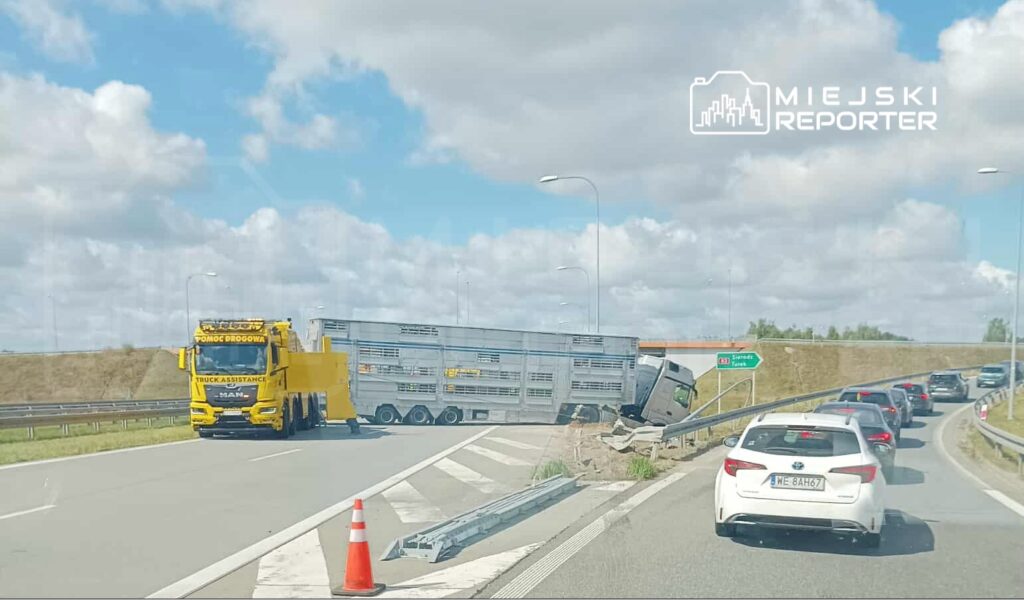 Żółty pojazd pomocy drogowej asystujący przy przewróconej ciężarówce na drodze, z utrudnionym ruchem samochodowym.