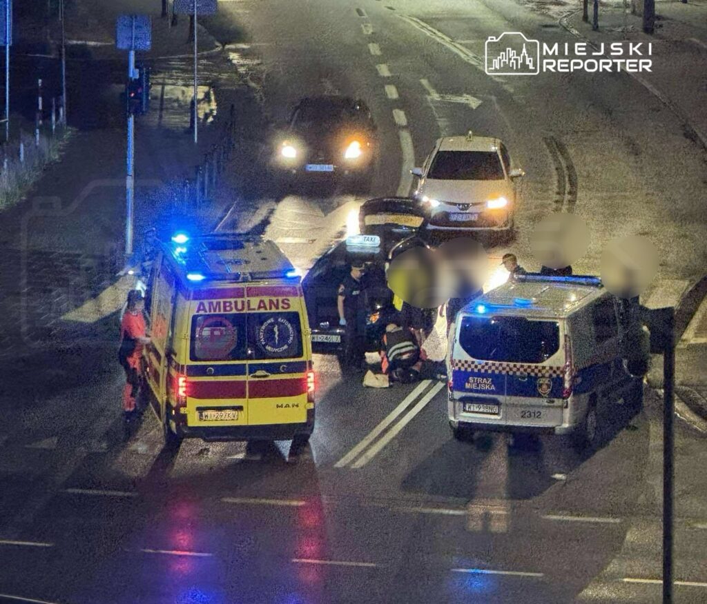 Na jezdni zatrzymane są pojazdy, a ratownicy medyczni udzielają pomocy poszkodowanemu w nocy.