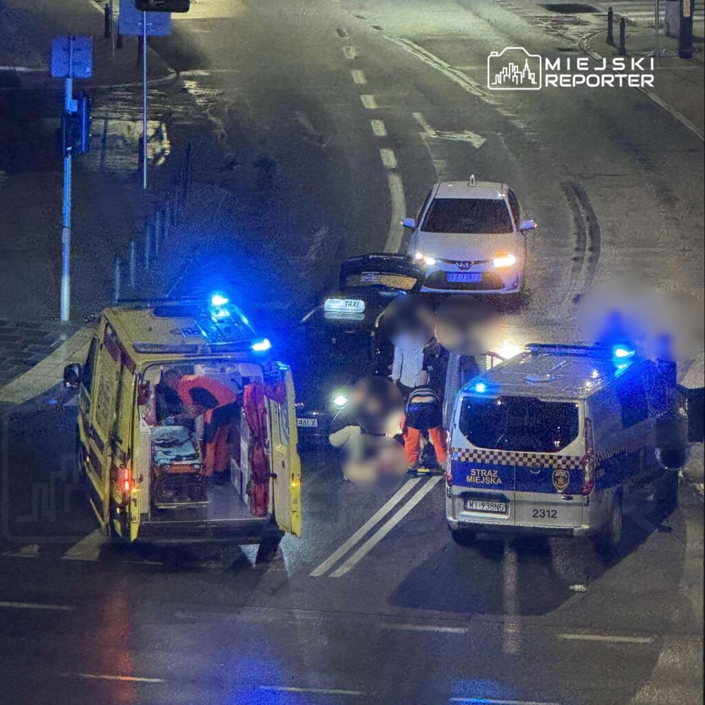 Zespół ratownictwa medycznego udziela pomocy poszkodowanemu na jezdni, obok zaparkowanego taxi i radiowozu.