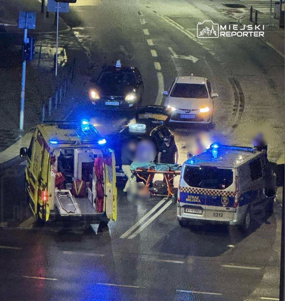 Ambulans z włączonymi sygnałami świetlnymi stoi na ulicy, a ratownicy przenoszą poszkodowanego na noszach.