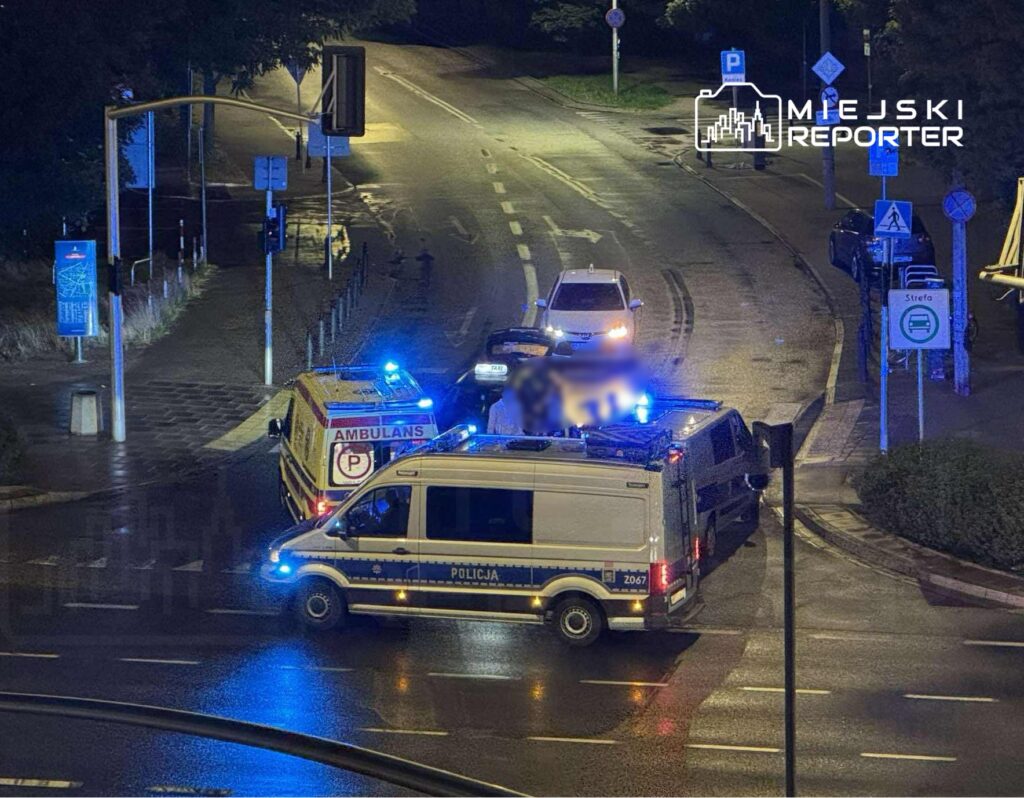 Na skrzyżowaniu ulic stoją pojazdy służb ratunkowych, w tym ambulans i radiowóz policji, otoczone niebieskimi światłami.