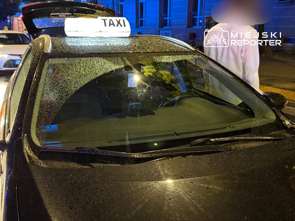 Taksi z podświetloną tablicą "TAXI" stoi na mokrej nawierzchni, w tle widać oświetlone budynki.