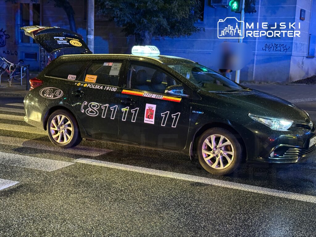 Czarny taksówka z otwartą klapą bagażnika stoi na pasach w oświetlonym miejskim otoczeniu nocą.