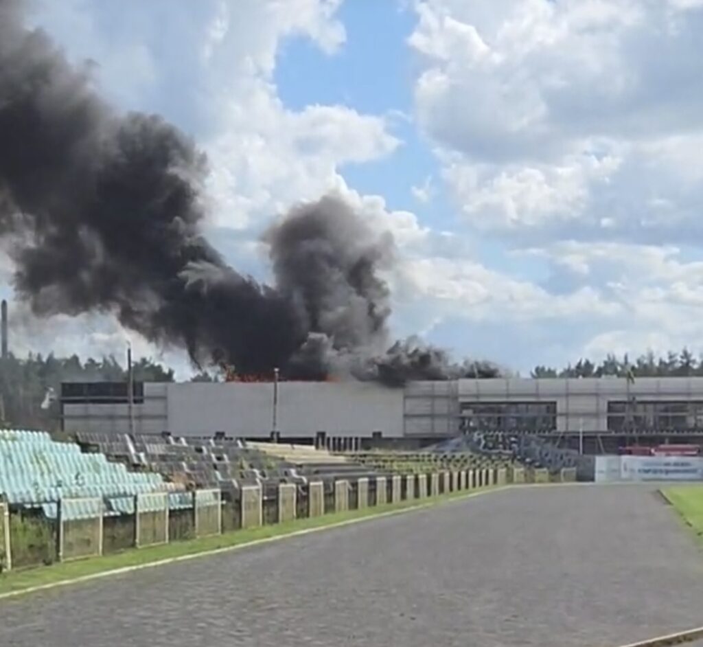 Czarny dym unoszący się z dachu nowoczesnego budynku w otoczeniu stadionu, w tle błękitne niebo i chmury.