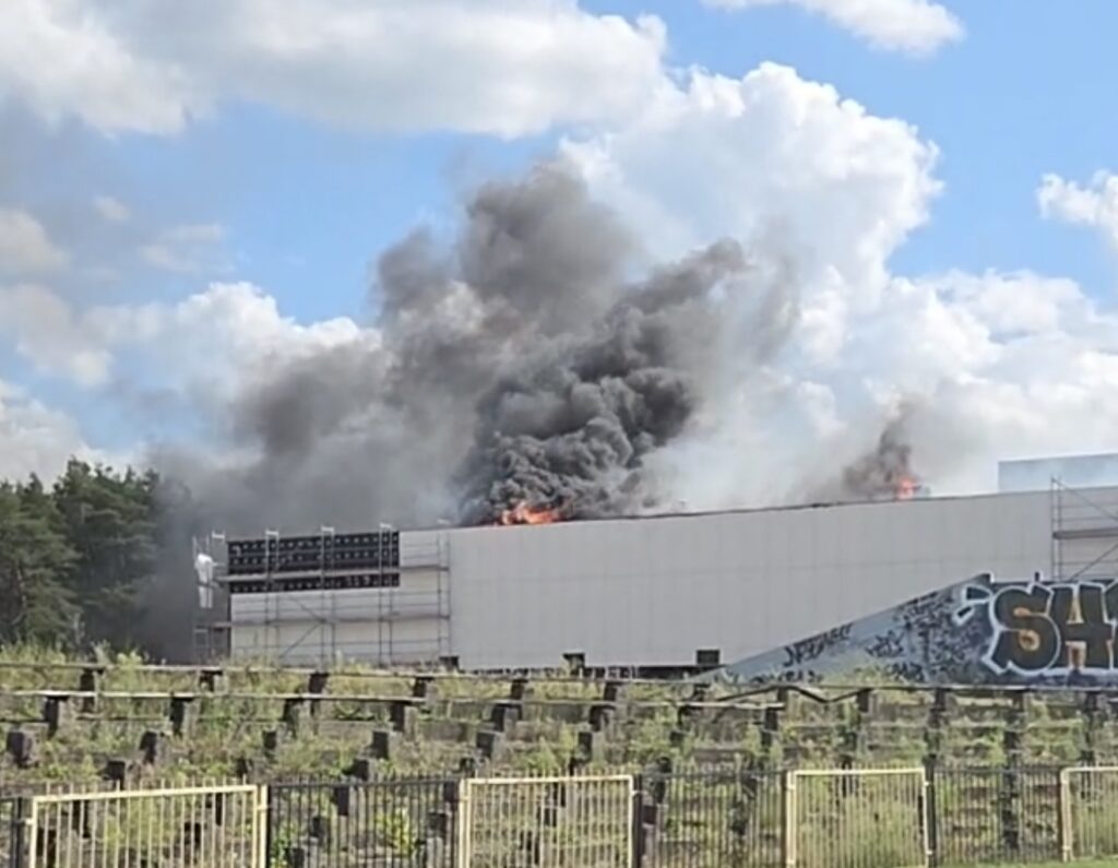 Czarne kłęby dymu i płomienie wydobywające się z dachu nowoczesnego budynku w otoczeniu zieleni.