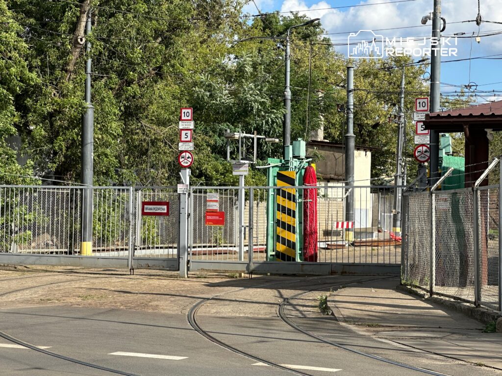 Brama wjazdowa z czerwonymi i żółtymi oznaczeniami, otoczona torami tramwajowymi i zielenią drzew.