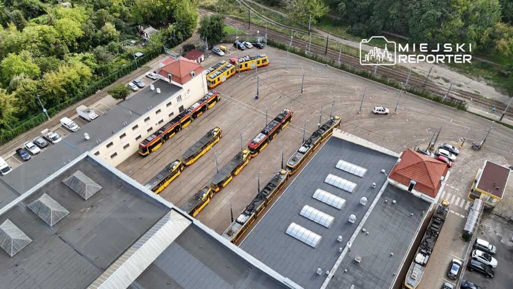 Tramwaje w żółto-czarnych barwach stoją na pętli w zajezdni, otoczone zielenią i parkingiem.