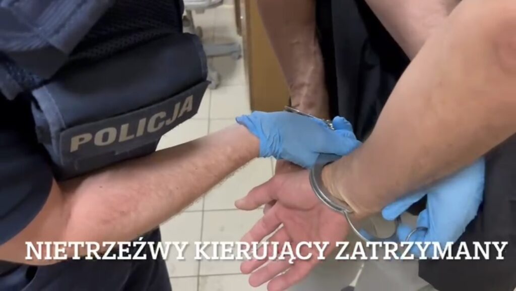 Policjant w mundurze zakłada kajdanki nietrzeźwemu kierującemu w pomieszczeniu z jasnymi ścianami.