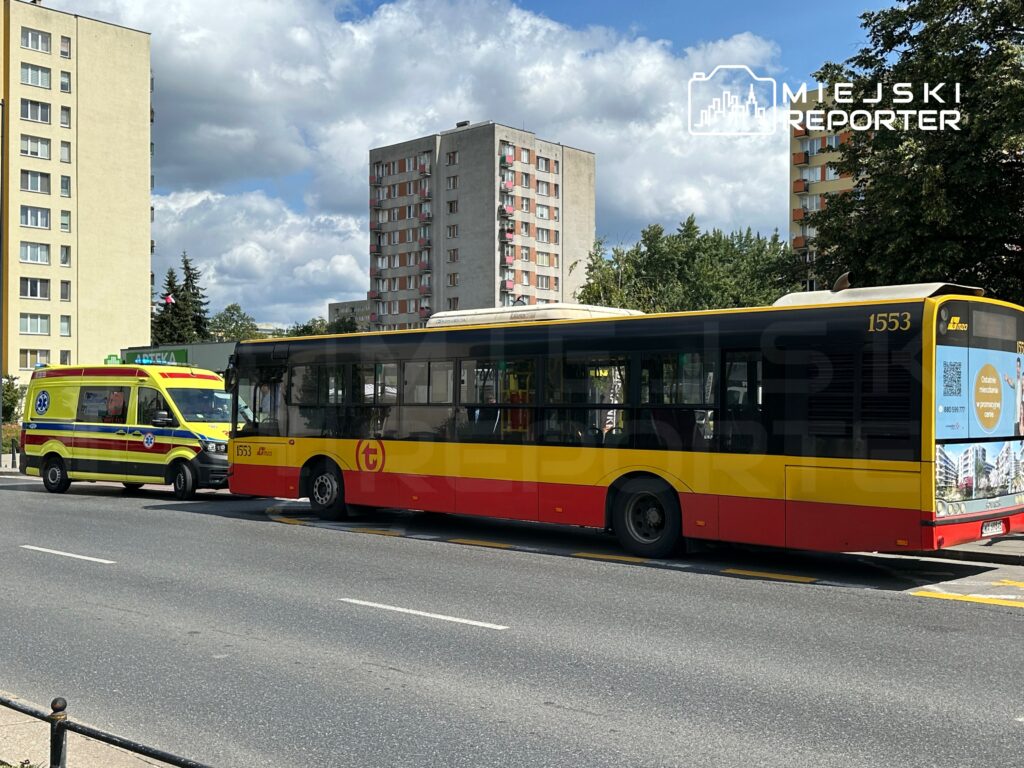 Karetka pogotowia zatrzymana obok miejskiego autobusu na ruchliwej ulicy, w tle bloki mieszkalne i niebo z chmurami.