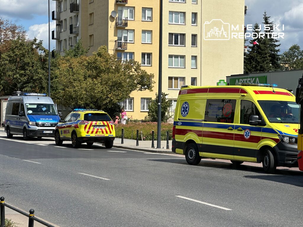 Radiowóz policji i karetka pogotowia stoją na ulicy przed blokiem mieszkalnym. W tle widoczni przechodnie.