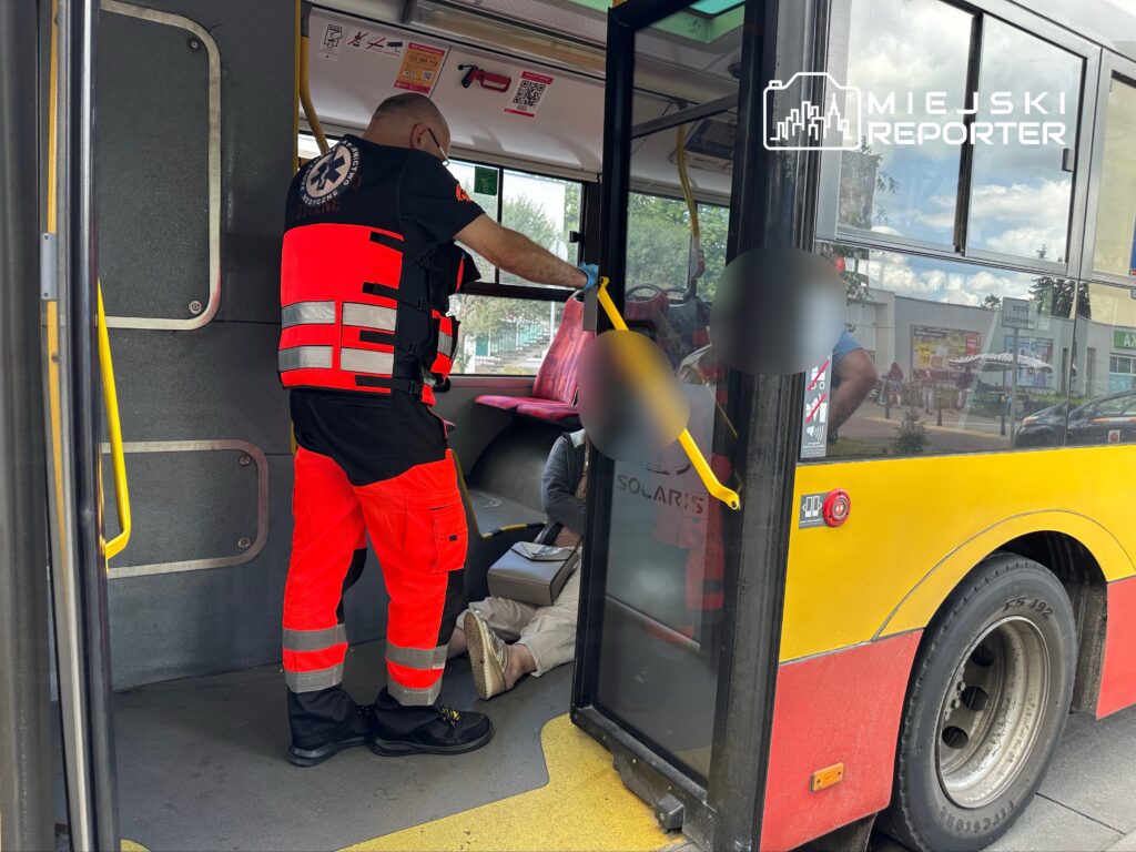 Ratownik medyczny w odblaskowej kamizelce udziela pomocy osobie siedzącej na podłodze autobusu miejskiego.