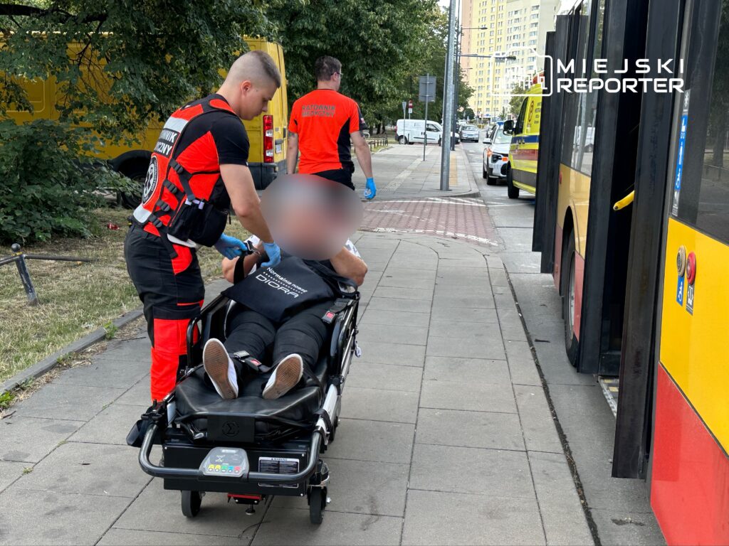 Ratownik medyczny udziela pomocy mężczyźnie leżącemu na noszach przy przystanku autobusowym.