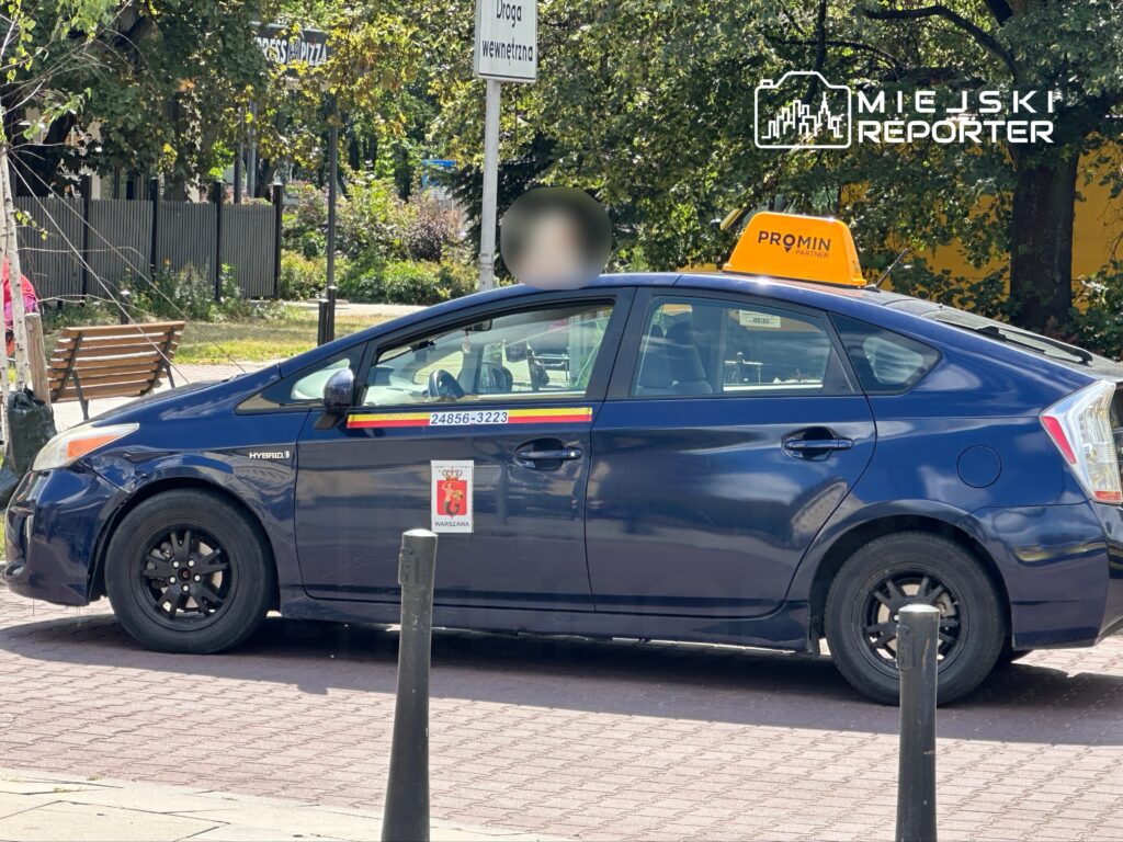 Taksi hybrydowe z żółtym oznaczeniem "PROMIN" zaparkowane na ulicy, w tle widać zieloną roślinność i ławkę.