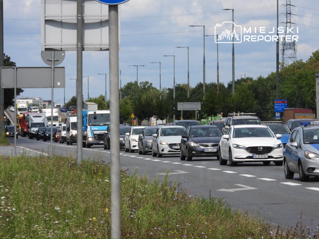 Ruch samochodowy na zatłoczonej drodze, z wieloma pojazdami stojącymi w kolejce, w otoczeniu zieleni i znaków drogowych.