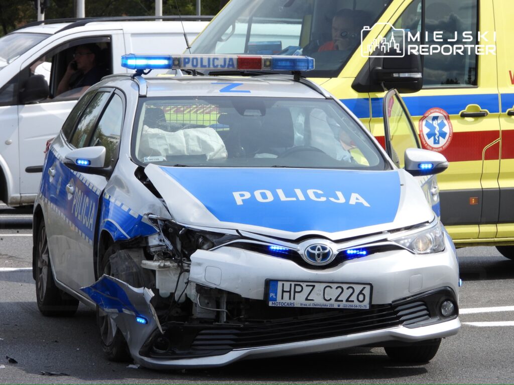 Uszkodzony radiowóz policyjny z niebieskimi światłami staje na drodze obok ambulansu, w tle widoczny kierowca w białym samochodzie.