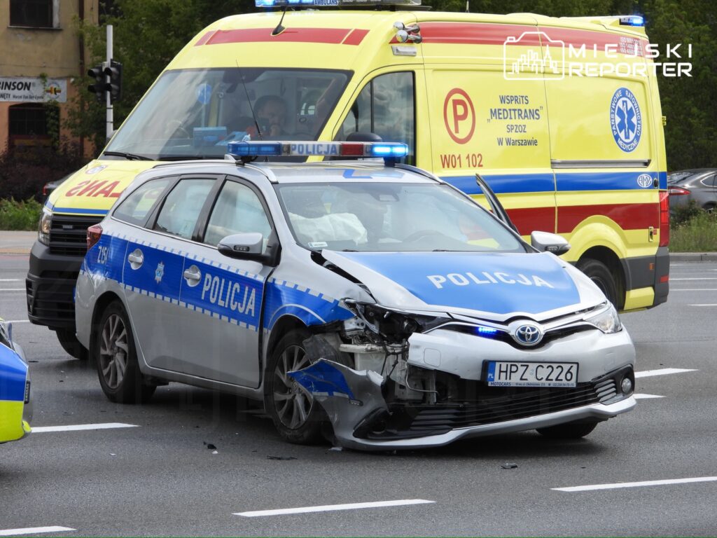 Policyjny radiowóz z uszkodzoną przednią częścią stoi na skrzyżowaniu, obok widać ambulans.