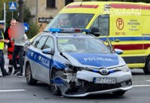 Policyjny radiowóz z uszkodzoną przednią częścią stoi na jezdni, obok funkcjonariusze rozmawiają z osobą poszkodowaną. W tle widać ambulans.