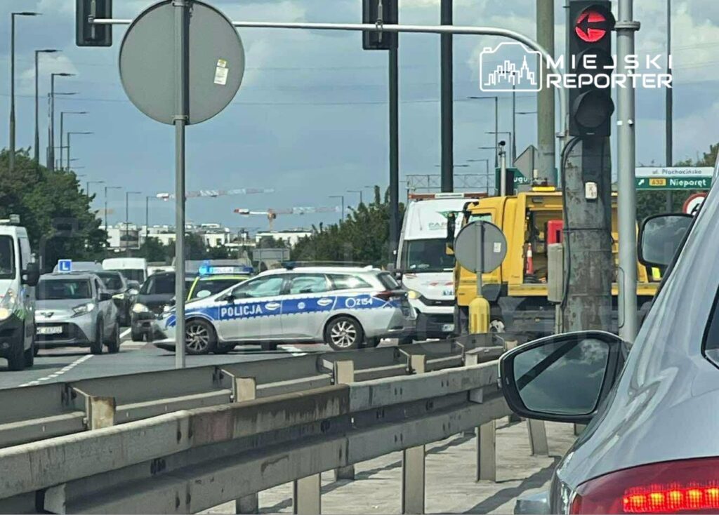 Ruch uliczny z policją kierującą pojazdami na skrzyżowaniu, w tle widoczne budowy i sygnalizacja świetlna.