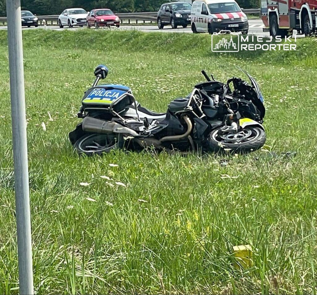 Zniszczony motocykl policyjny leży na trawie obok drogi, w tle widoczne pojazdy osobowe.