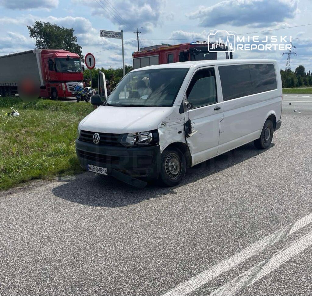 Uszkodzony biały Volkswagen stoi na poboczu drogi, obok czerwonego ciężarowego pojazdu i wozu strażackiego.