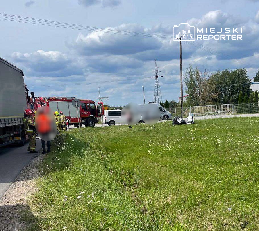 Grupa strażaków w kaskach pracuje przy drodze, obok stojącego samochodu dostawczego i wozu strażackiego.