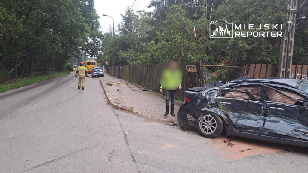 Mężczyzna w odblaskowej kamizelce stoi obok rozbitego samochodu na drodze, obok widać policyjny radiowóz.