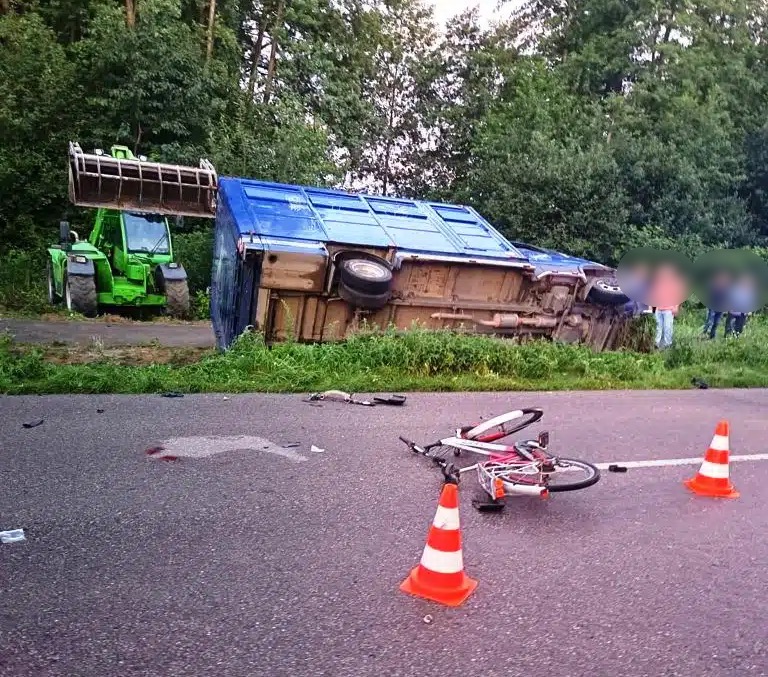 Przewrócony niebieski kontener leży na boku przy drodze, obok stoi zielony traktor, a na jezdni leży uszkodzony rower.