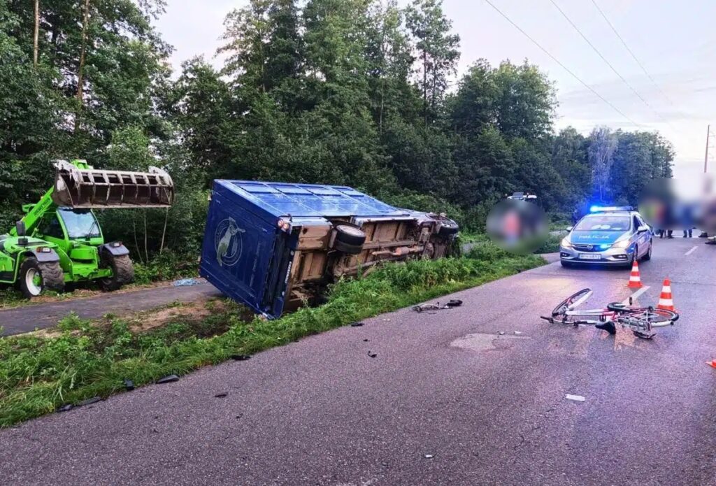 Na drodze leży przewrócony niebieski kontener, obok stoi zielony ciągnik z podnośnikiem. W tle widać radiowóz policyjny z włączonymi sygnałami.