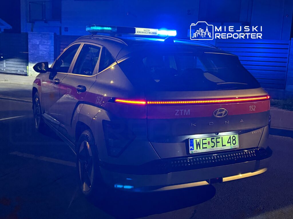 Radiowóz elektryczny marki Hyundai Kona z włączonymi niebieskimi sygnalizatorami stoi na ulicy w nocy.