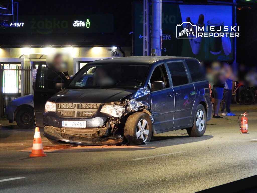 Uszkodzony minivan z wgniecioną przednią częścią stoi na jezdni, obok pomarańczowy trójkąt ostrzegawczy. W tle widoczny sklep Żabka.