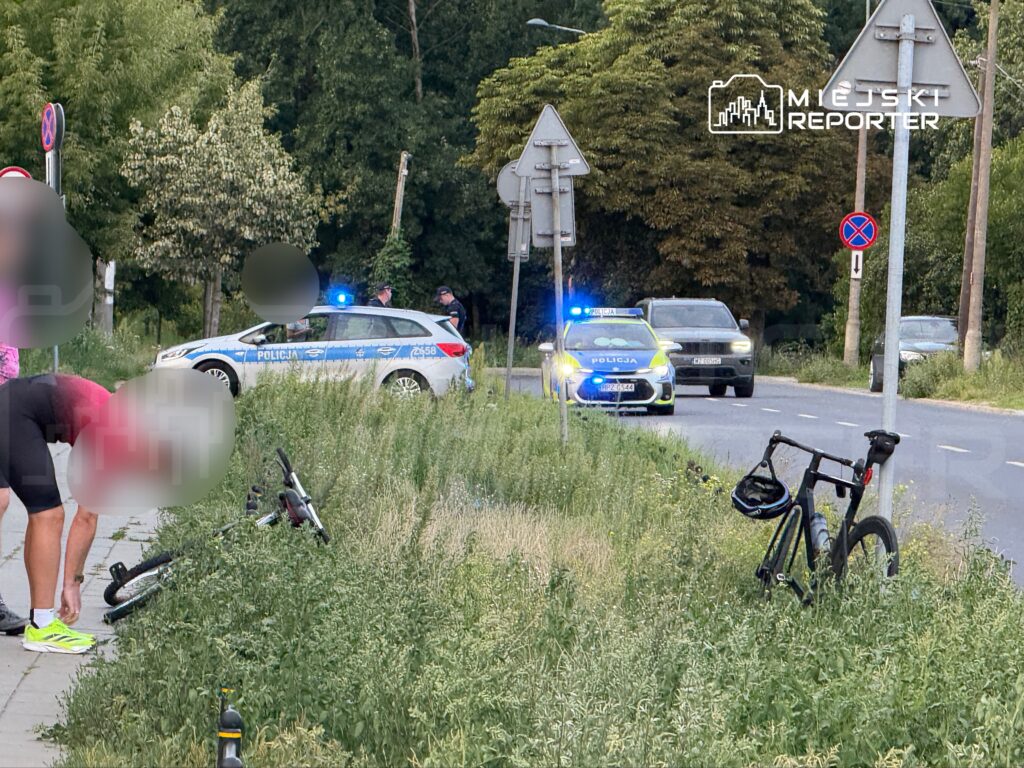 Mężczyzna w różowej koszulce schyla się przy rowerze na poboczu drogi, obok policyjny radiowóz z włączonymi sygnałami.