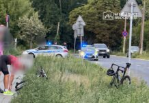 Mężczyzna w różowej koszulce schyla się przy rowerze na poboczu drogi, obok policyjny radiowóz z włączonymi sygnałami.