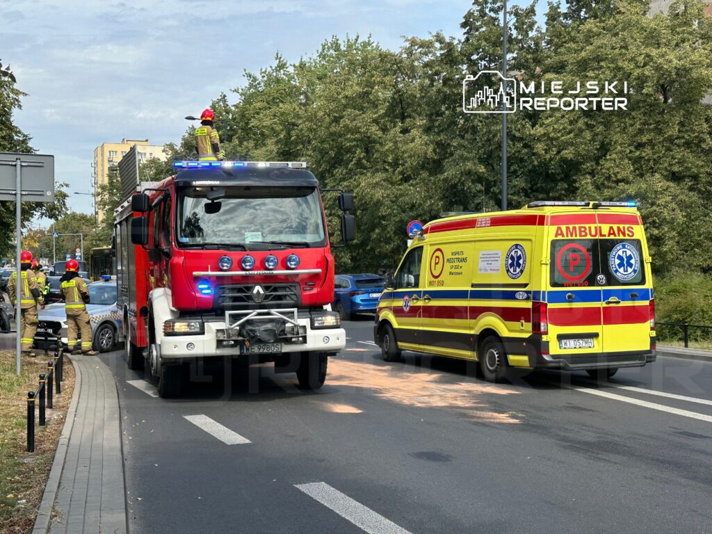 Strażacy w hełmach pracują przy wozie strażackim, obok ambulans z włączonymi światłami na ruchliwej ulicy.