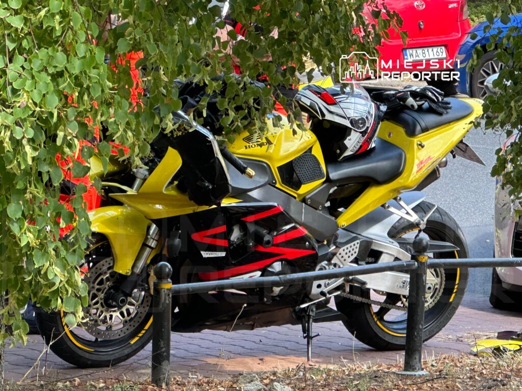 Motocykl w kolorze żółtym z czarnymi akcentami zaparkowany w cieniu drzew, obok leżą kask i rękawice.