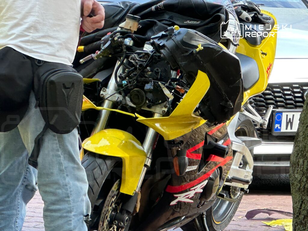 Mężczyzna w białej koszulce stojący obok uszkodzonego żółtego motocykla na chodniku.