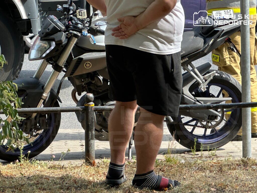 Mężczyzna w krótkich spodniach stoi z rękami za plecami obok motocykla marki Yamaha na chodniku.