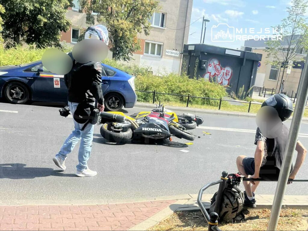 Motocykl leży na jezdni, a obok stoi mężczyzna w kurtce skórzanej, obserwujący sytuację. W tle widać zaparkowany samochód.