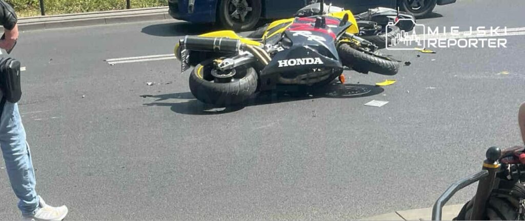 Motocykl leży na boku na jezdni, obok stoi mężczyzna z torbą, a w tle widać inne pojazdy.