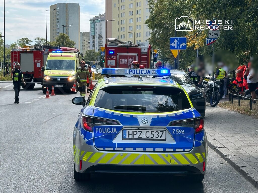 Radiowóz policyjny z włączonymi sygnałami świetlnymi stoi na ulicy, obok pracują strażacy i karetka pogotowia.