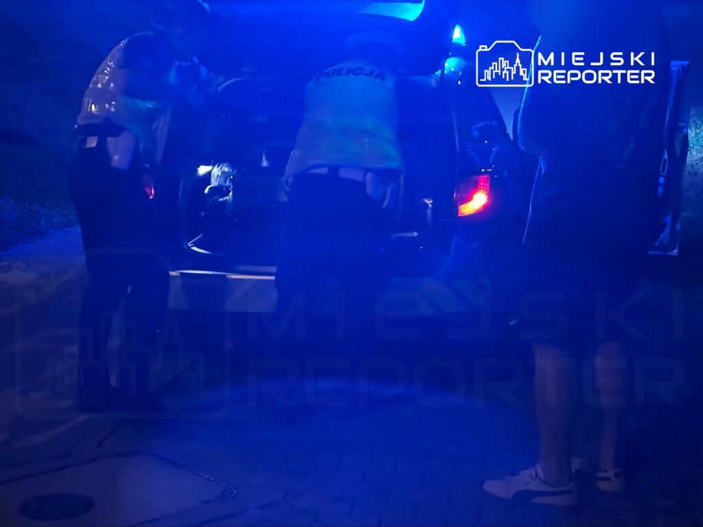 Dwaj funkcjonariusze policji przeszukują bagażnik samochodu w nocy, w otoczeniu niebieskiego światła.