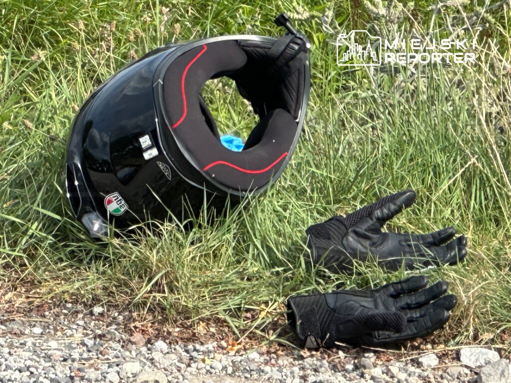 Czarny kask motocyklowy i rękawice leżą na trawie obok drogi, otoczone kamieniami i zielenią.